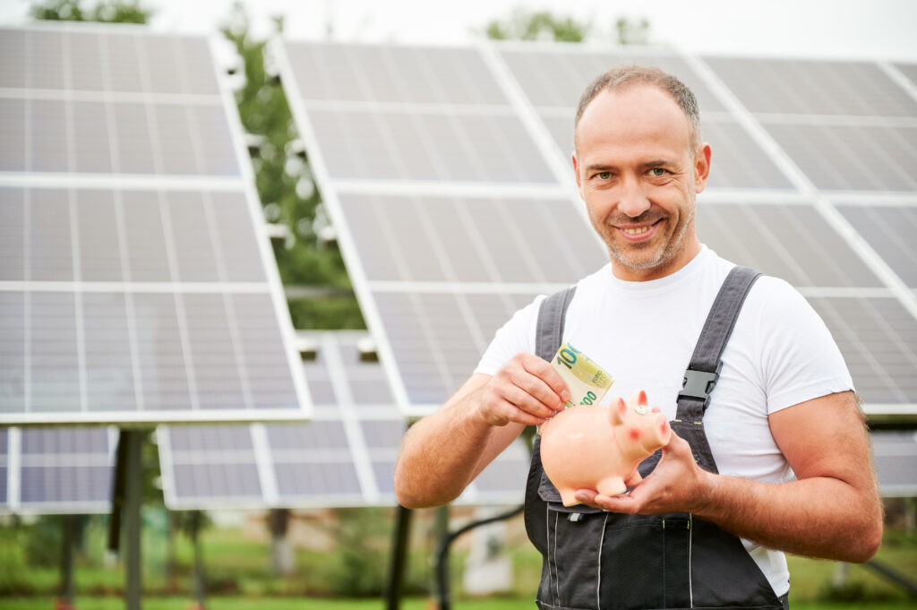 Mann mit Sparschwein vor Photovoltaik Anlage